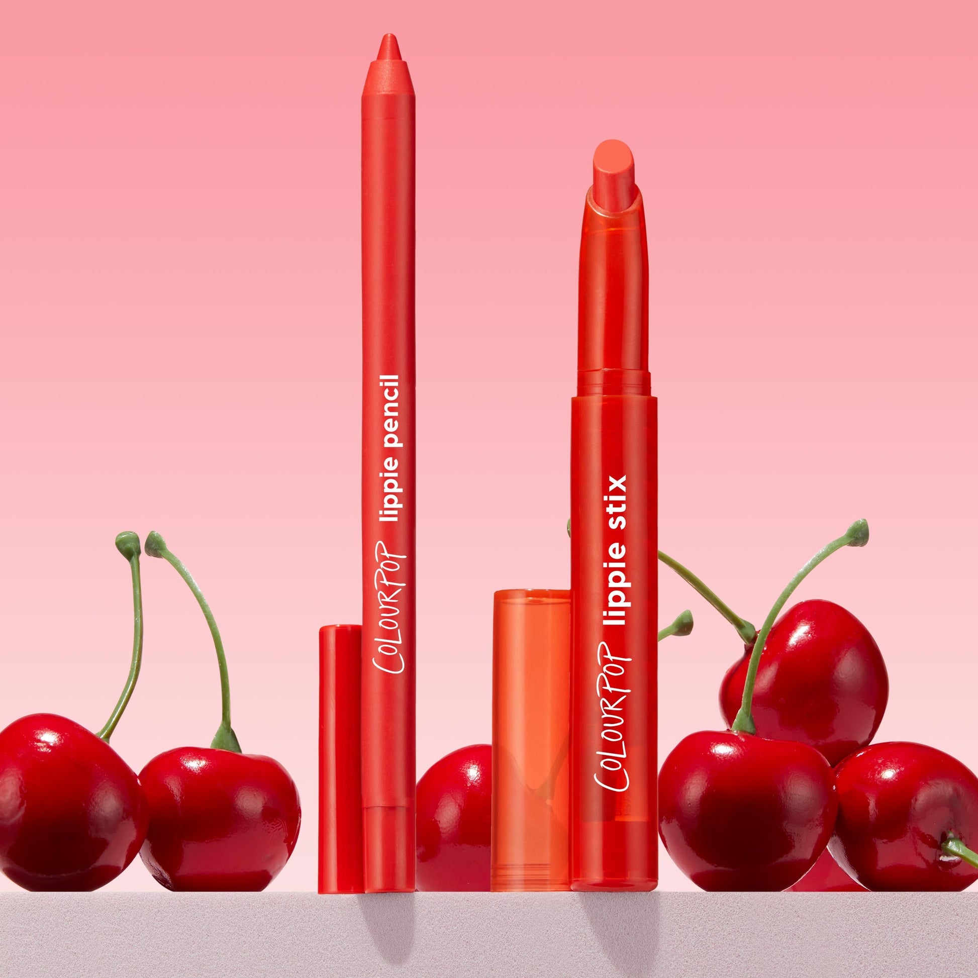 ColourPop Chi Chi Lippie Stix and ColourPop Lippie Pencil stand uncapped to show vivid red tips, set among glossy cherries on a pink gradient background, highlighting the bright cherry-red lip duo for bold, defined color.