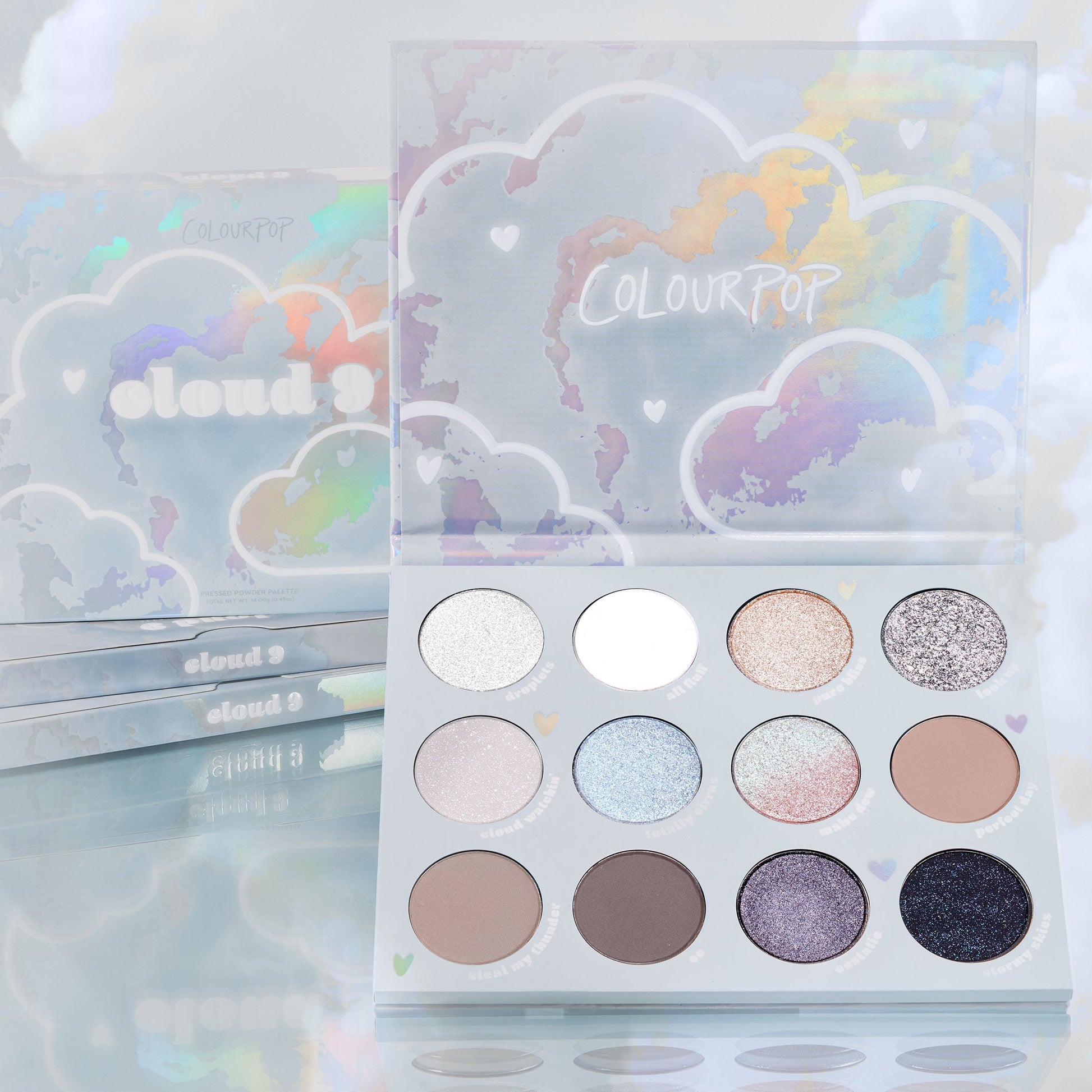 Cloud 9 ColourPop Shadow Palette opens to reveal 12 cool-toned neutrals and iridescent shimmers - from icy silver and matte white to taupe, lilac, and deep navy - set in holographic cloud packaging with stacked palettes and pastel clouds behind.
