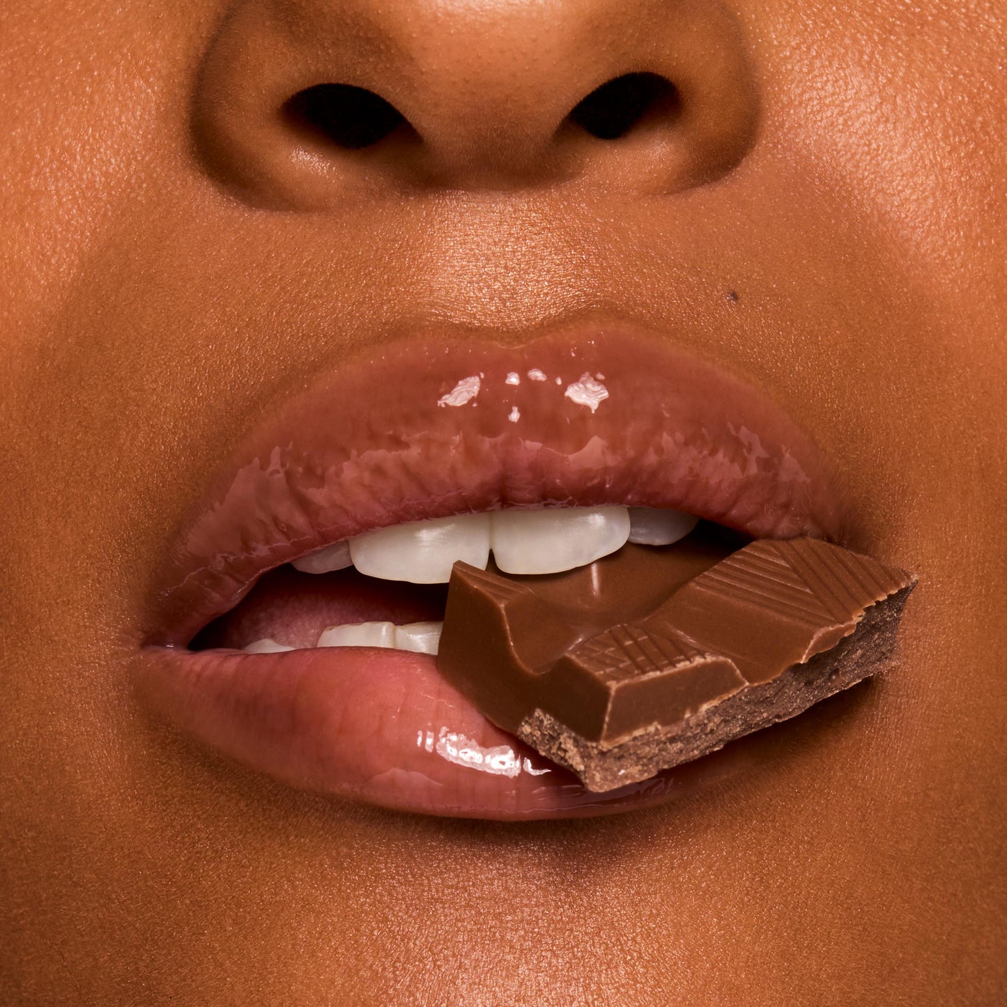 Glossy, cocoa-tinted lips wearing ColourPop Fresh Kiss Glossy Balm in Chocolate Milkshake hold a square of milk chocolate between teeth in a macro close-up, showcasing high-shine, cushy hydration and a creamy nude tint.